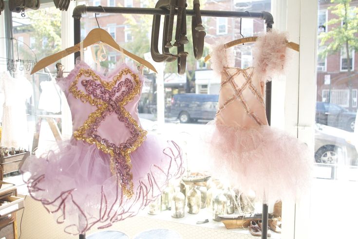 two tutus are displayed in a store window, with one being a pale pink with gold and purple sequins and the other a more feathery pink tutu with golden details. the window is decorated with various glass bottles and vases. the store is likely a dance or clothing store.
