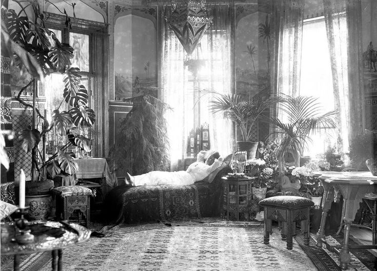 a black and white photograph depicting the interior of a sunlit room in a home, possibly from the early 1900s. a woman is reclining on a daybed, surrounded by numerous potted plants including palm trees and monstera deliciosa. the room is decorated in an eclectic style, featuring moroccaninspired furniture, ornate rugs, and wall decorations or murals. the overall atmosphere is peaceful and serene. the image suggests wealth and a relaxed lifestyle.