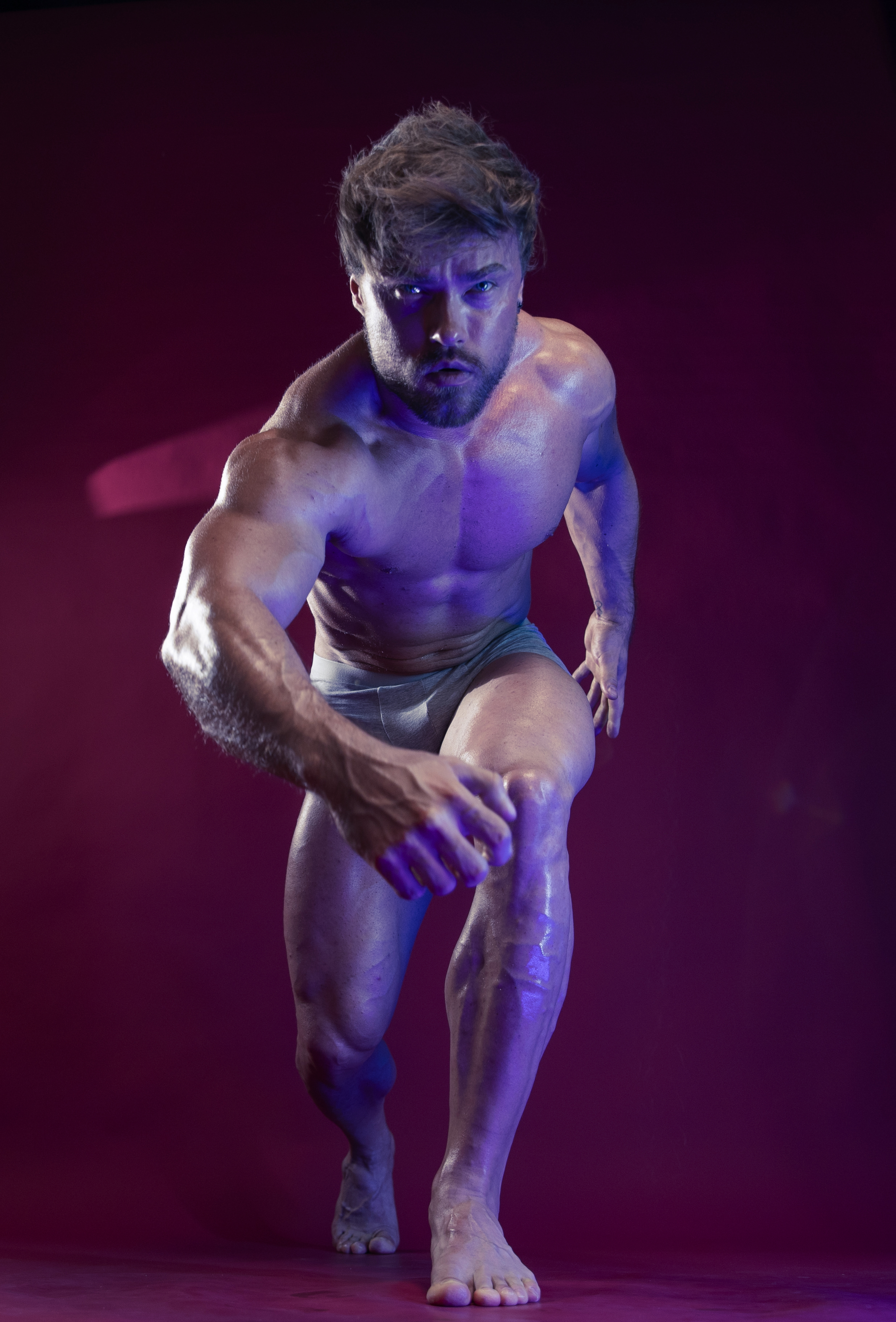 a muscular man in a dynamic pose, possibly a bodybuilder or athlete. he is shirtless and appears oiled or sweaty, highlighting his physique and visible veins. the dramatic lighting, with purplish hues, creates a sense of intensity and determination, further emphasized by his focused expression and forward motion. the background is a solid purple color.