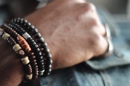 a closeup shot of a person's wrist wearing a stack of bracelets. the bracelets are made of black wood and brown wood beads.  a silver band with a small stone is on one of the bracelets.  the person's hand is resting on a piece of blue denim fabric.  the background is out of focus.