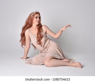 a beautiful woman with long red hair is wearing a white dress and a crown. she is sitting on a white background and looks elegant and graceful. she is posing for a photo shoot and has a very natural and relaxed pose. she looks like a goddess.