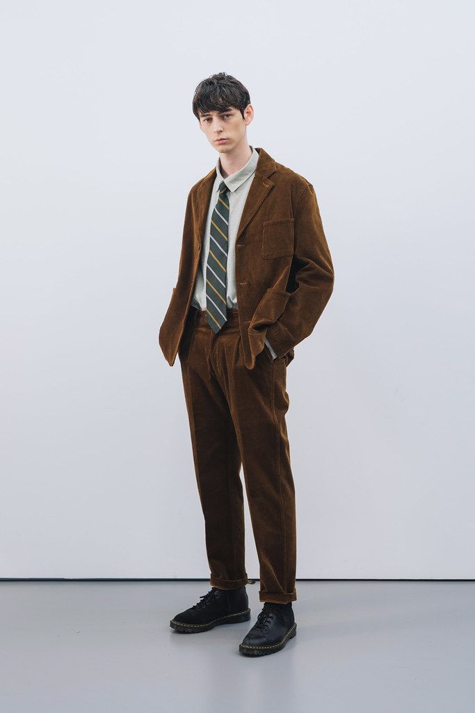 fulllength studio shot of a male model wearing a brown corduroy suit. the suit consists of a corduroy jacket and matching corduroy pants.  he is wearing a lightcolored shirt and a striped tie.  he has dark hair and is wearing dark brown or black shoes. the background is a plain light gray. the overall style is smart casual.