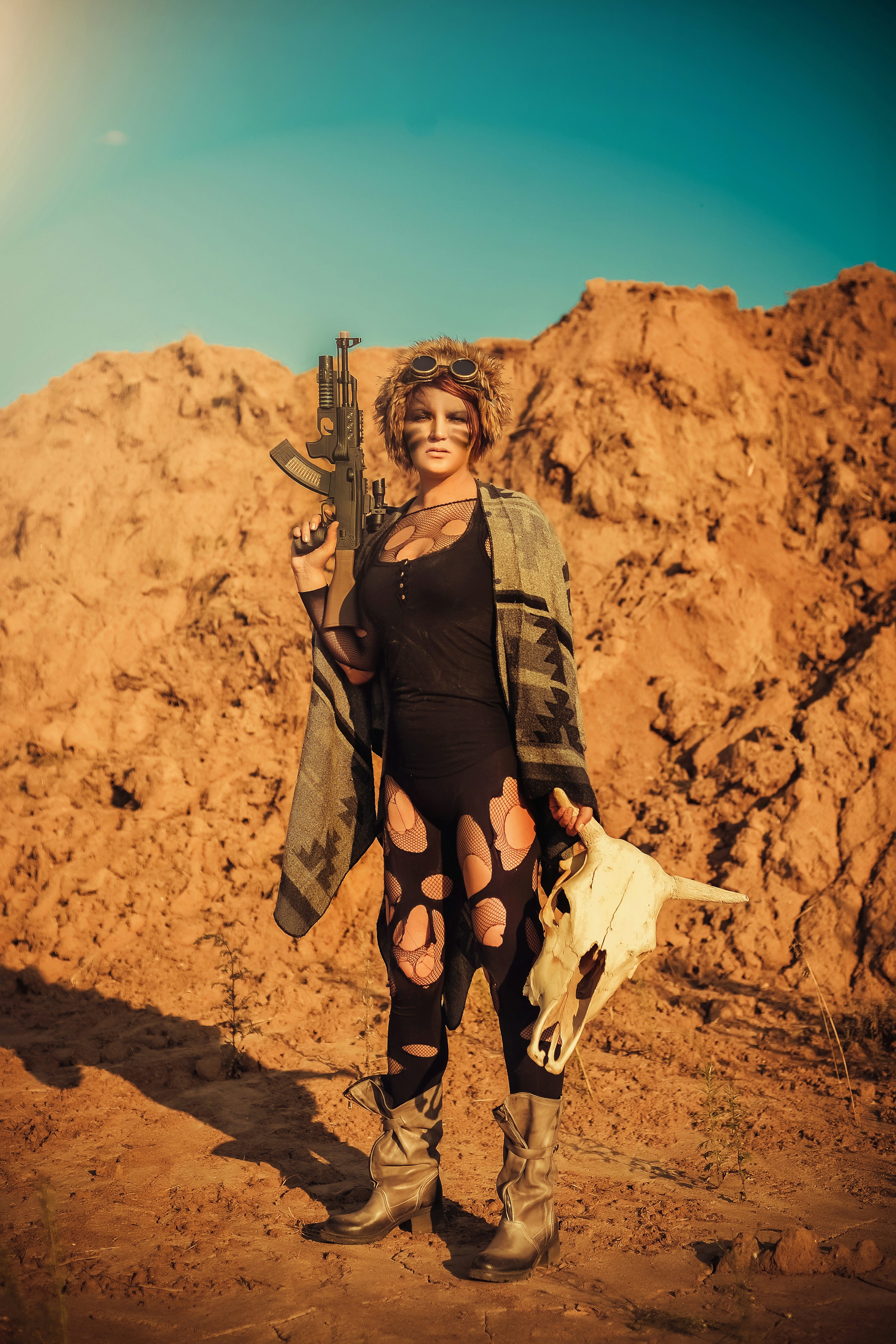 a full shot of a female warrior in a postapocalyptic setting. she stands in a barren, sandy landscape, holding an ak47 rifle and a cow skull. her attire consists of ripped leggings, boots, a poncho, and goggles. her face is painted, enhancing the fierce and survivalist look. the image has a sepiatoned, vintage feel, suggesting a dramatic and atmospheric setting.