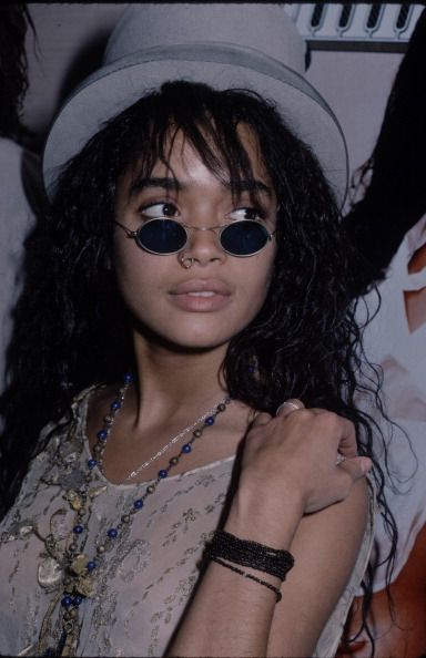 close up portrait of a young woman with long, curly dark hair wearing round sunglasses, a hat, a white dress and jewelry. she is looking at the camera with a serious expression. the image is likely from the 90s.
