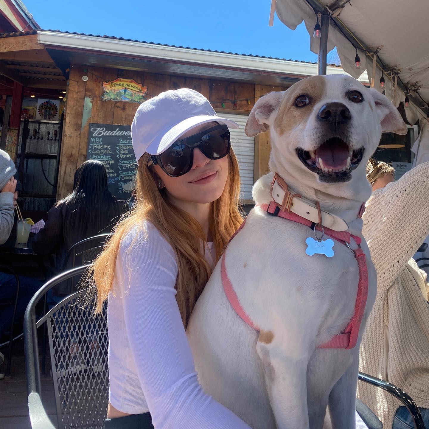 a young woman with long reddishblonde hair is sitting outdoors at a patio restaurant, happily holding a white dog in her lap.  she's wearing a white baseball cap and sunglasses. the dog is wearing a pink harness and has a calm and friendly expression. the setting appears to be a casual, sunny outdoor cafe or bar.  a menu board is visible in the background.