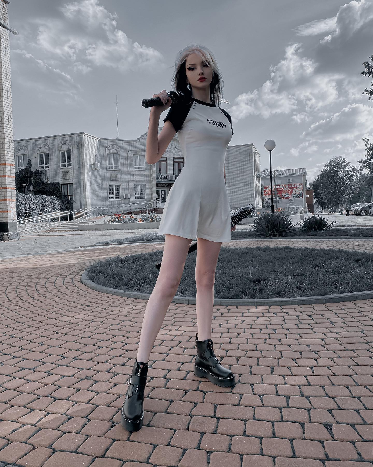 a fullbody shot of a young woman, possibly a model, standing on a cobblestone street in an urban setting.  she's wearing a short, white dress with black sleeves, and black chunky boots. she is holding what looks like a microphone or similar object. the overall aesthetic is dark, grunge, and slightly monochrome with a cool color palette. the background is slightly blurred but shows buildings and a cloudy sky.  the woman is posing, and the image has a fashionoriented feel.