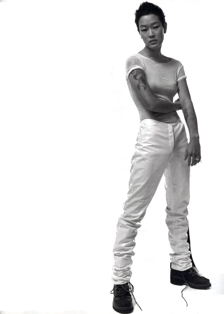 a black and white photo features a woman with short hair and tattoos, wearing white pants and a tshirt, standing in boots.