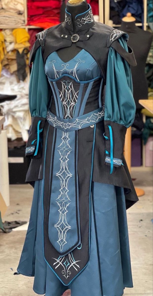 a mannequin is dressed in a teal and black fantasystyle dress with silver embellishments. the dress features a fitted bodice, puffed sleeves, and a long skirt. the bodice and skirt are accented with black panels and silver embroidery. the dress is completed with a short cape and collar.