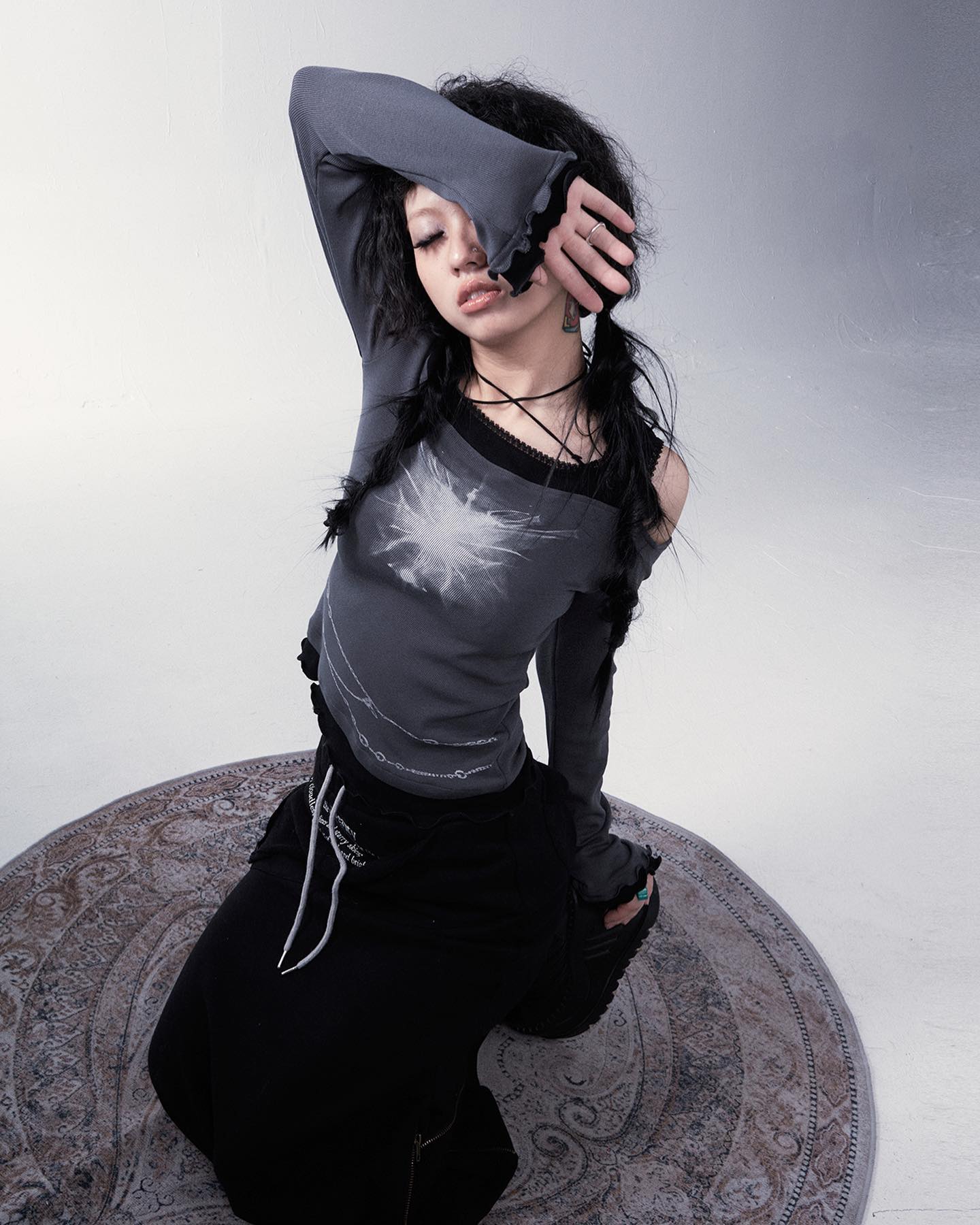 a young woman is posing for a fashion photoshoot. she is wearing a grey longsleeved top with a unique graphic design and a black skirt.  her dark hair is styled loosely, partially covering her face. she is sitting on a patterned rug on a white floor against a light grey backdrop. the overall style is dark, edgy and alternative. the image is artistically styled with a focus on the model's clothing and pose.