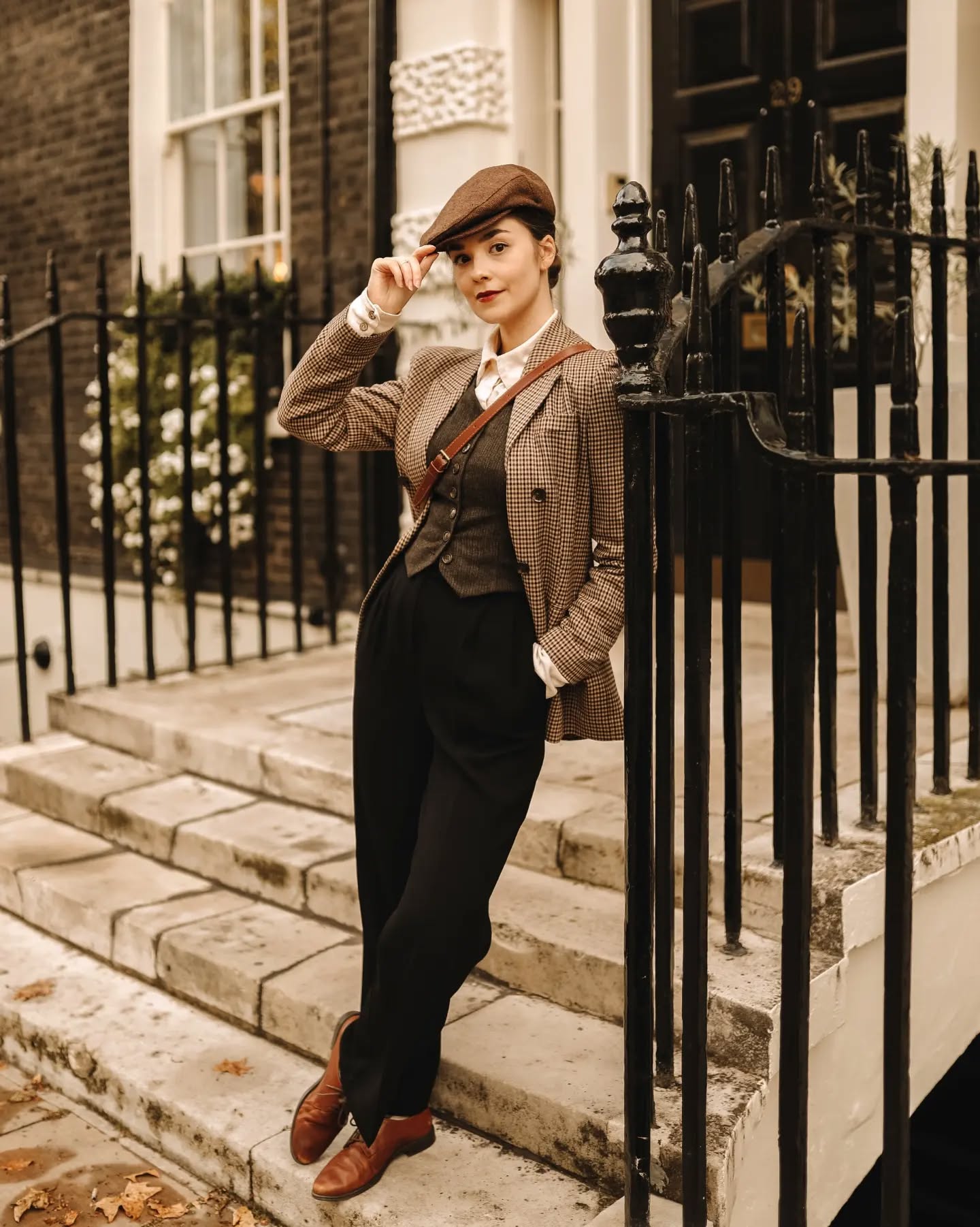 a woman in a vintageinspired outfit stands on a set of steps outside a london building. she's wearing a brown tweed jacket, a darkcolored waistcoat, black trousers, brown oxfords, and a newsboy cap.  she carries a brown satchel bag across her body. the setting appears to be autumnal.  the photo has a vintage or retro aesthetic.