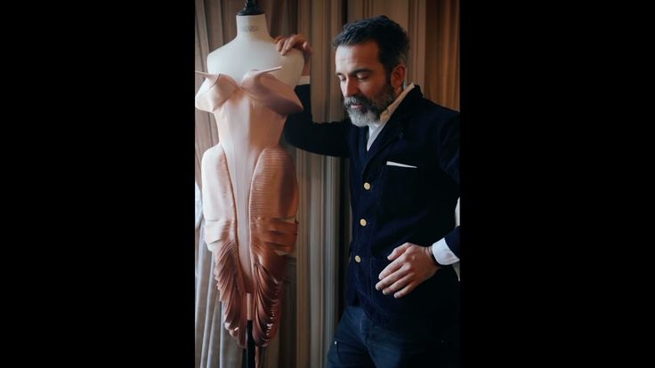 a man with a beard, identified as a fashion designer, stands next to a mannequin wearing a unique dress. the designer is wearing a suit. the scene appears to be set in a studio or workshop.