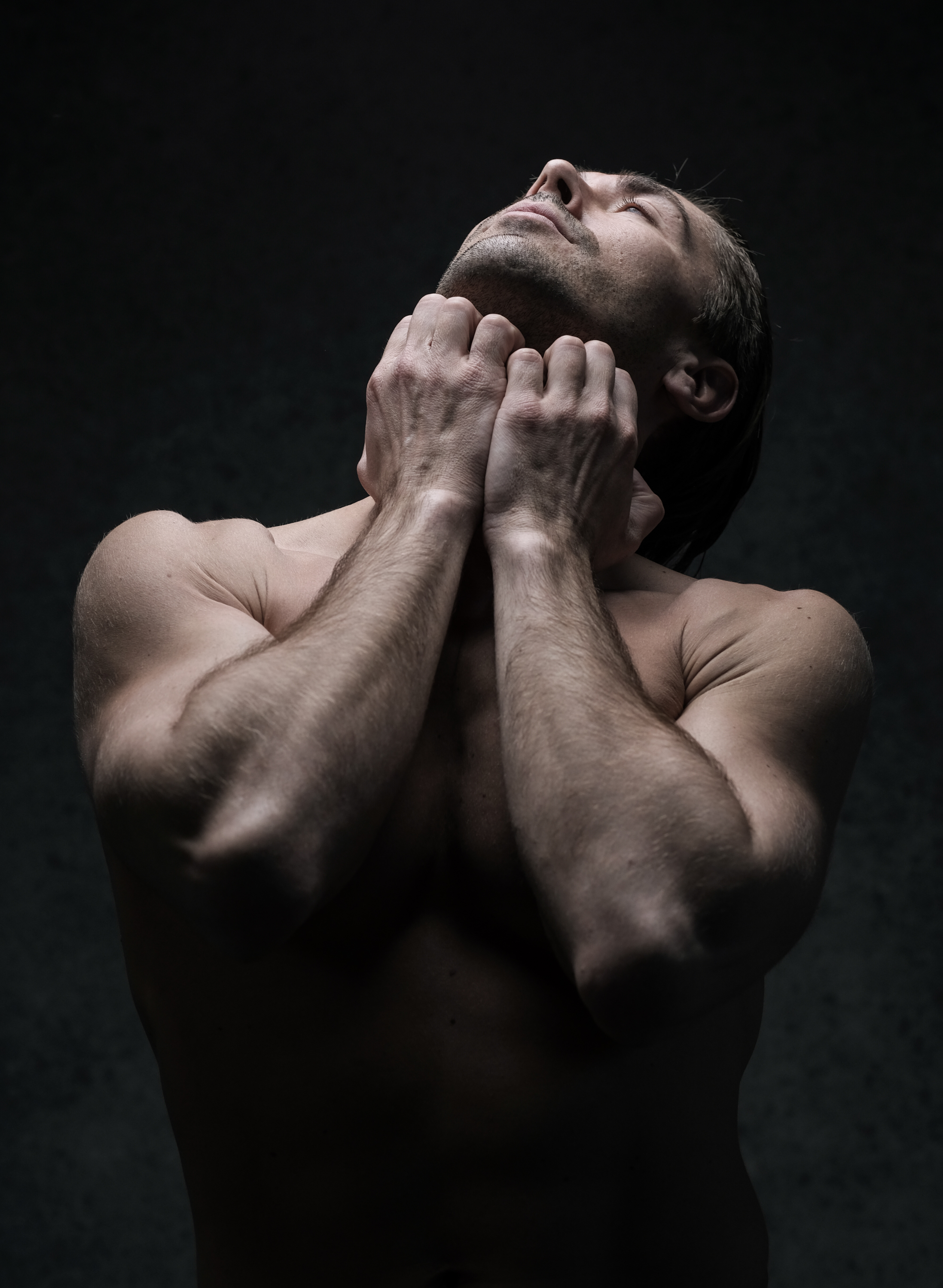 a shirtless muscular man stands with his head tilted back, eyes closed, and hands clasped around his neck in a dimly lit setting. the dramatic lighting emphasizes his physique.