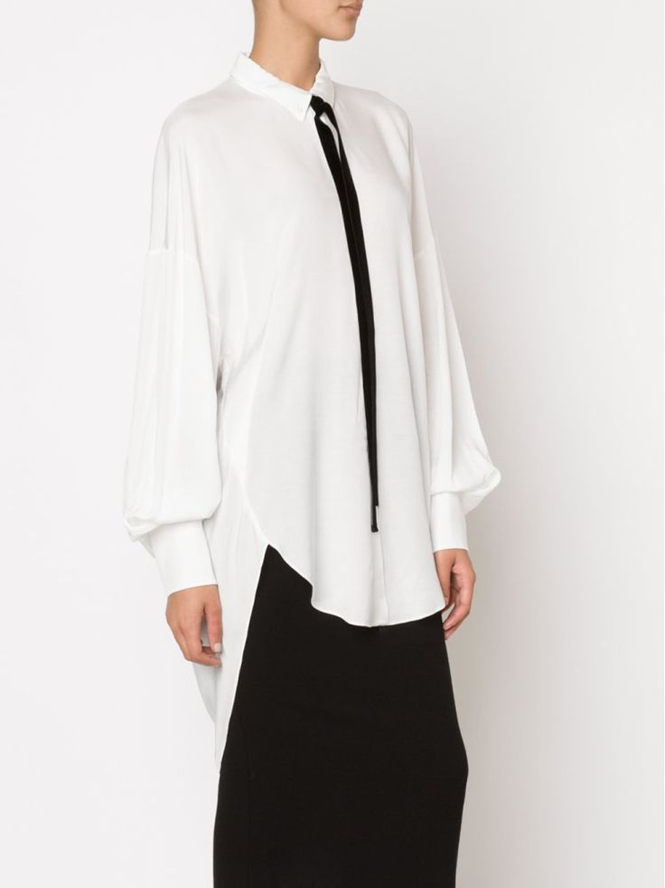 a woman is wearing a white longsleeved blouse with a black tie and a black skirt. she is standing in front of a white background. the blouse has a highlow hemline and loose sleeves with buttoned cuffs. the skirt is kneelength and fitted. the overall look is minimalist and chic.