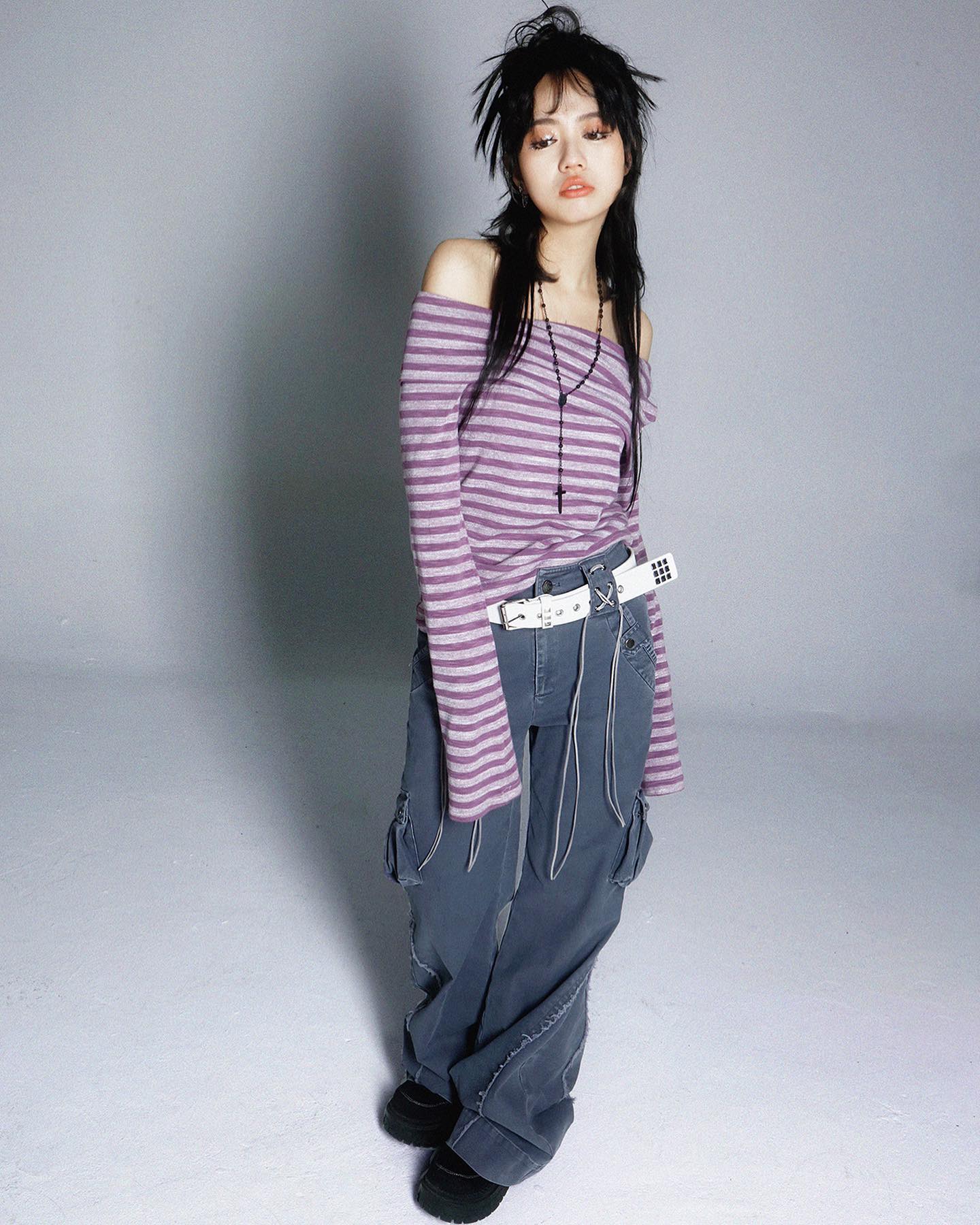 a full shot of a young woman posing for a fashion photograph.  she's wearing a purple and white striped offtheshoulder longsleeved shirt, grey cargo pants, a white belt with black accents, a black cross necklace, and darkcolored boots.  her dark hair is styled in a choppy, slightly messy fashion. she is standing on a plain light grey background. the overall style is casual yet edgy.