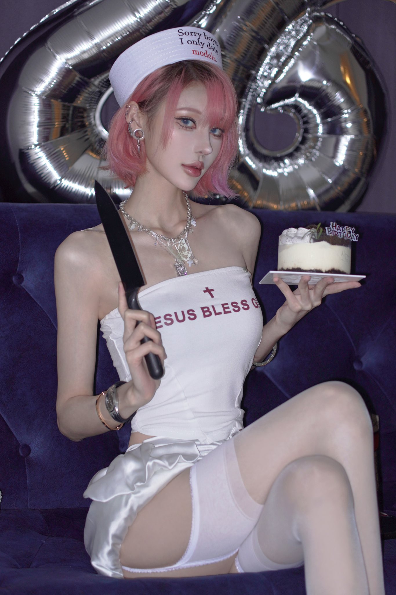 a young woman with short pink hair and a sailor hat is sitting on a purple couch. she is holding a knife in one hand and a birthday cake in the other. she is wearing a white dress with a "jesus bless" message and white stockings with a garter belt. there are silver balloons in the background.