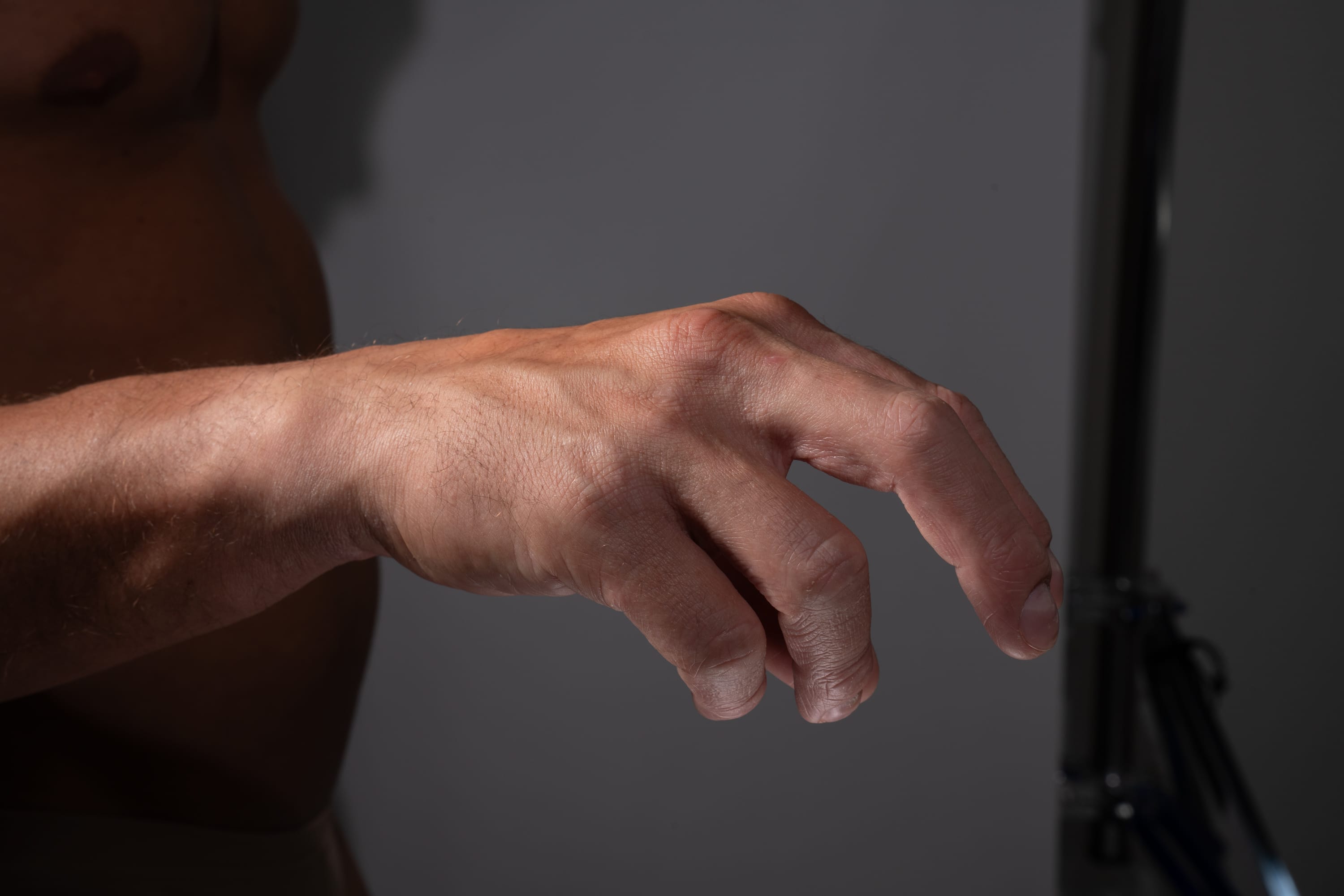 the image showcases a closeup of a man's hand and torso, illuminated by studio lighting against a neutral gray background. the perspective emphasizes the contours and details of his hand, highlighting its gesture and anatomy. the muted color palette contributes to a naturalistic feel.