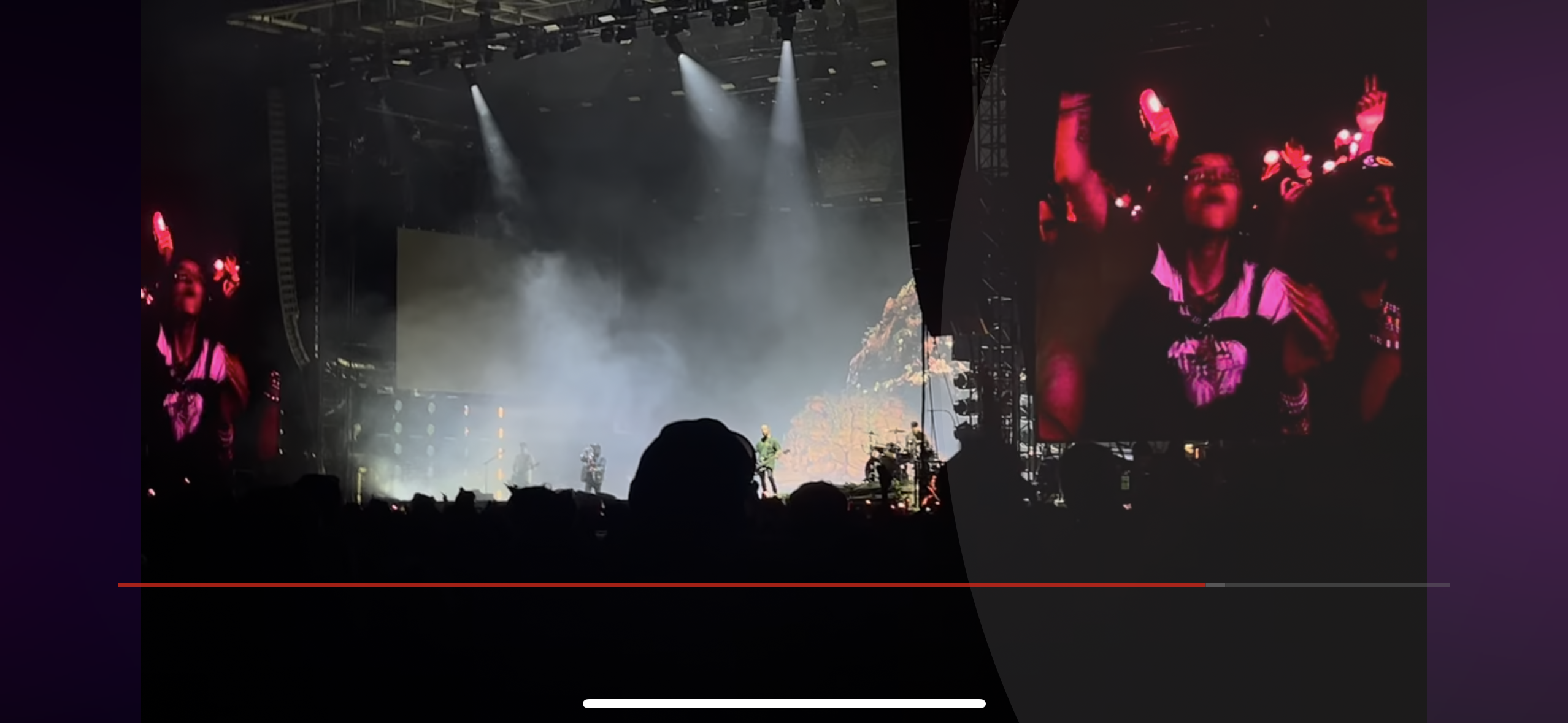 a concert scene at an indoor venue, a silhouette of a person is standing on stage with a mic stand, the stage is lit by spotlight and there is a projector screen behind the person and a crowd in the foreground is blurred, it's a night concert and there's a screen showing a person who looks excited and happy.