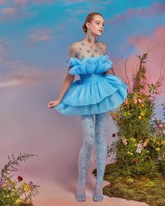a beautiful woman in a light blue tulle dress stands with a sweet smile on her face, posing in a whimsical garden setting. the dress is decorated with delicate floral details and creates a flowing, ethereal look. the model's soft makeup and wavy hair enhance the ethereal feeling of the image.