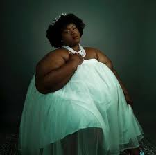 a black woman in a green formal dress is posing in a studio setting. she is sitting with her legs crossed and is looking at the camera with a confident smile. she has a lovely, elegant look.
