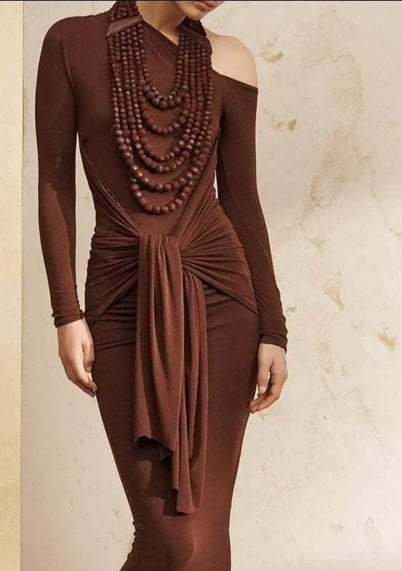 a woman in a brown oneshoulder dress poses elegantly in a studio setting. she wears a layered brown beaded necklace that contrasts with the fabric of her dress. the dress is simple, but stylish.