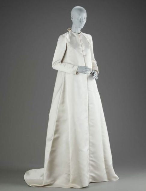 a fulllength studio shot showcases a minimalist wedding dress on a gray mannequin against a gray backdrop. the ivory satin gown features a long, flowing skirt and a simple, highnecked bodice. the design is elegant and understated, typical of 1960s haute couture. the dress hangs gracefully, emphasizing the fluidity of the fabric.  the overall aesthetic is clean and sophisticated.