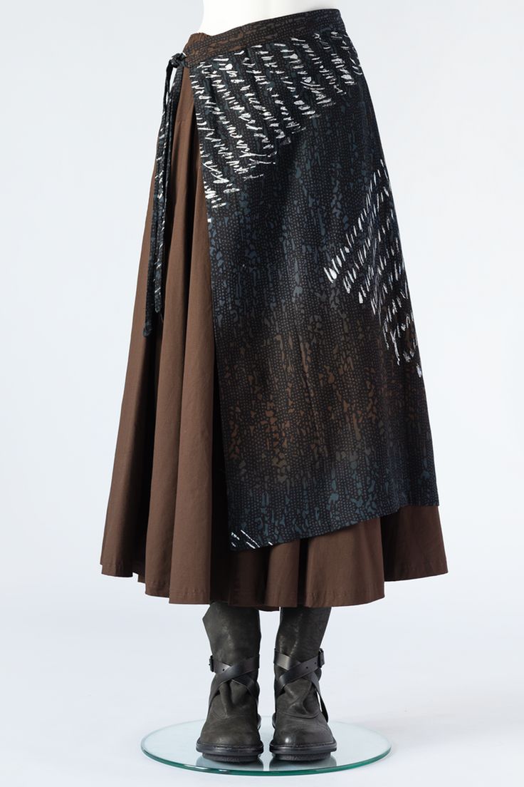 a full shot of a brown and black layered skirt on a mannequin. the skirt is composed of a brown midi skirt layered over a black patterned wraparound skirt. the pattern on the wraparound skirt is abstract with white and gray lines and shapes against a dark background. the mannequin is wearing dark brown ankle boots with buckles. the background is pure white.