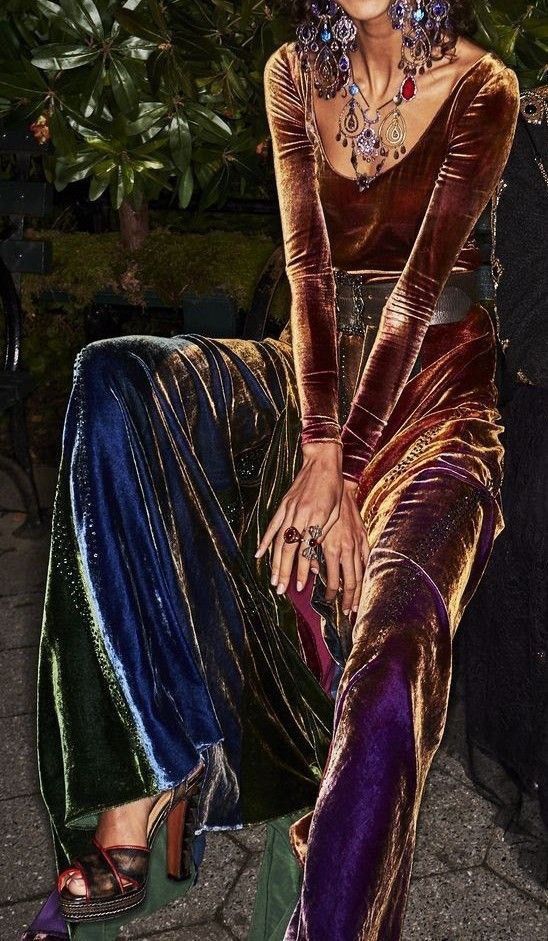 loseup shot of a woman wearing a stunning, multicolored velvet jumpsuit. the jumpsuit features varying shades of deep green, blue, purple, and gold velvet, creating a rich and luxurious look. the woman is adorned with statement jewelry, including layered necklaces, large earrings, and rings, enhancing the overall opulent style. she's also wearing highheeled sandals. the background is blurred, suggesting an outdoor setting with lush greenery. the image portrays a bohemian and sophisticated aesthetic. the overall feel is elegant and captivating.