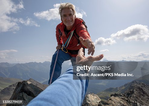 a man reaches out to help someone from the top of a mountain. the man is wearing a red jacket and blue pants, and he has a rope attached to his safety harness. the sky is blue with some clouds, and the sun is shining. the mountain range is visible in the background. the person being helped is wearing a blue shirt.