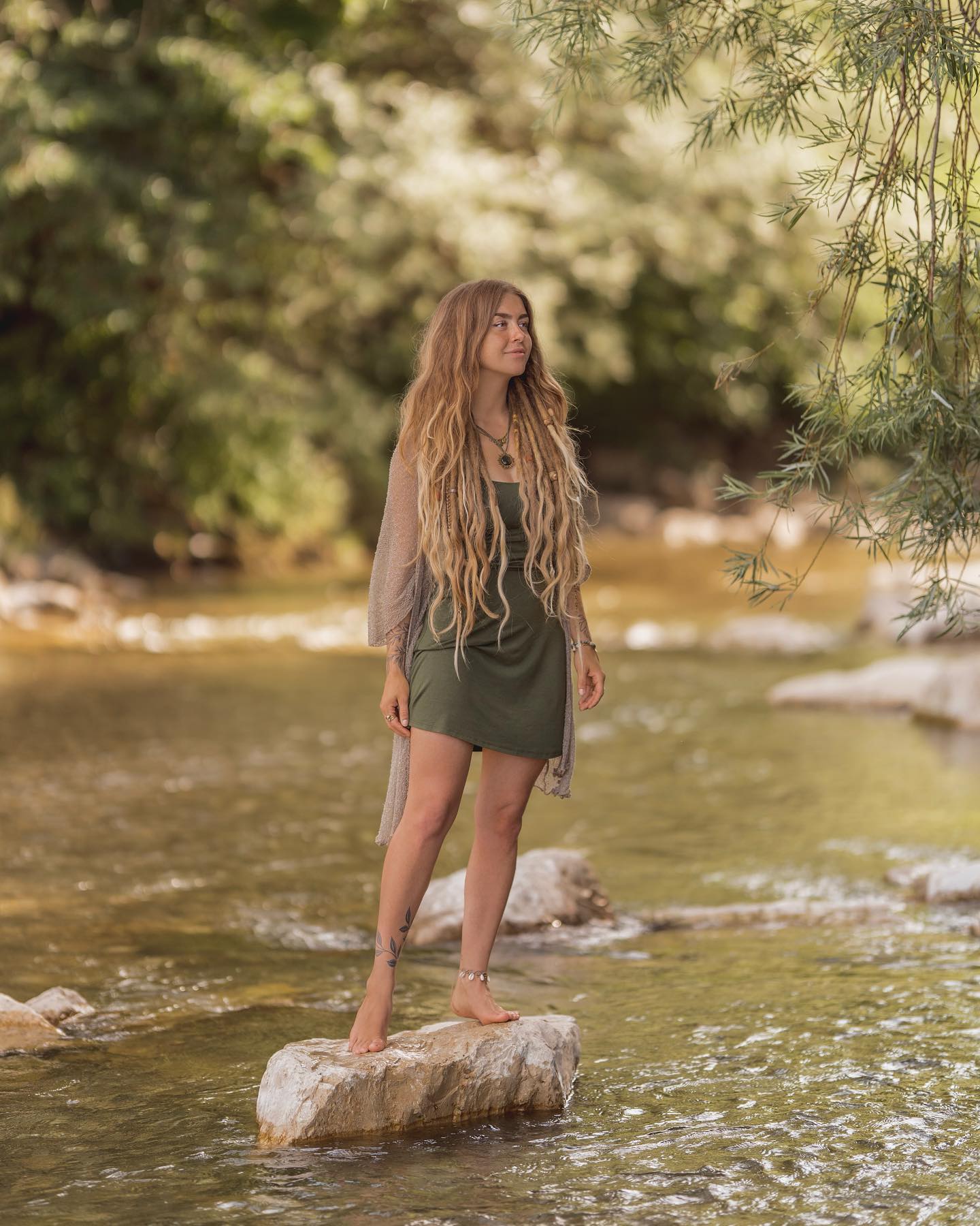 a young woman with long blonde dreadlocks stands barefoot on a rock in a shallow river. she is wearing a green dress and a beige cardigan. the setting is a peaceful and tranquil natural environment with lush greenery and sunlight filtering through the trees. the overall mood is serene and spiritual.