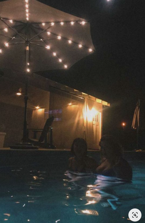 two women are swimming in a pool at night. the pool is lit by string lights strung across an open umbrella and warm lighting from inside a nearby home. the water is dark and reflective. the scene is relaxed and suggests a social gathering.