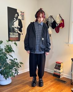 a man in a black jacket, black pants, and black boots is standing in a room. he is wearing a plaid shirt and headphones. the man is looking at the camera and smiling. there are books on a shelf in the background.