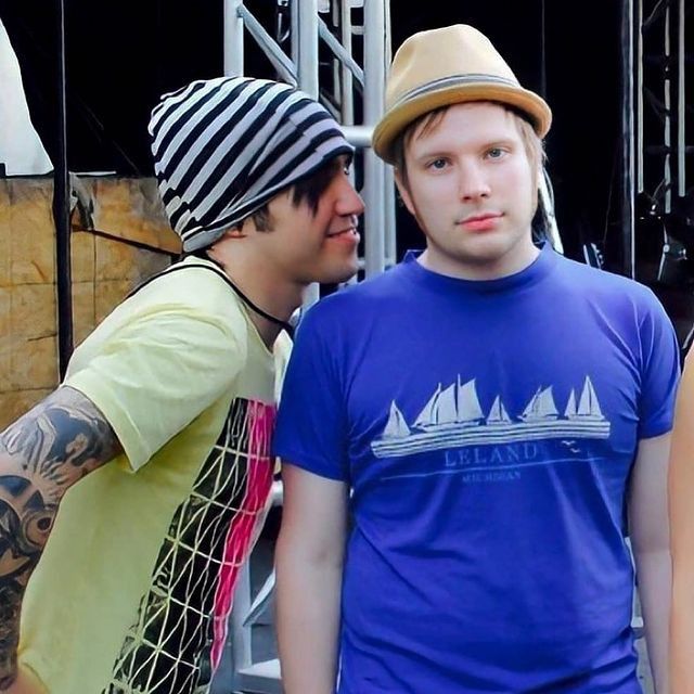 a photo of patrick stump and pete wentz of fall out boy. patrick is wearing a blue tshirt and a beige fedora, smiling at the camera. pete is wearing a yellow and pink tshirt and a black and white striped beanie, smiling and looking at patrick. they appear to be backstage at a concert.
