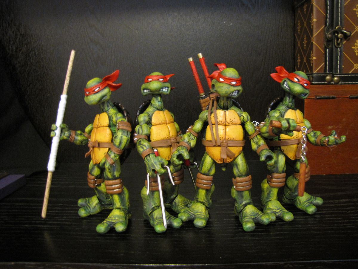 the image shows four teenage mutant ninja turtles action figures lined up sidebyside. each turtle has a red mask and carries a different weapon: a bo staff, sai, katanas, and nunchucks. the figures are standing on a dark surface with a dark background. a wooden box with a brown and tan pattern is visible to the right.