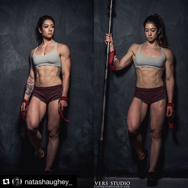 two women athletes pose with a barbell. they are both incredibly ripped and muscular, wearing sports bras and shorts. the women are staring intensely at the camera, as if they are ready to compete.  they stand on a dark background in a studio setting.