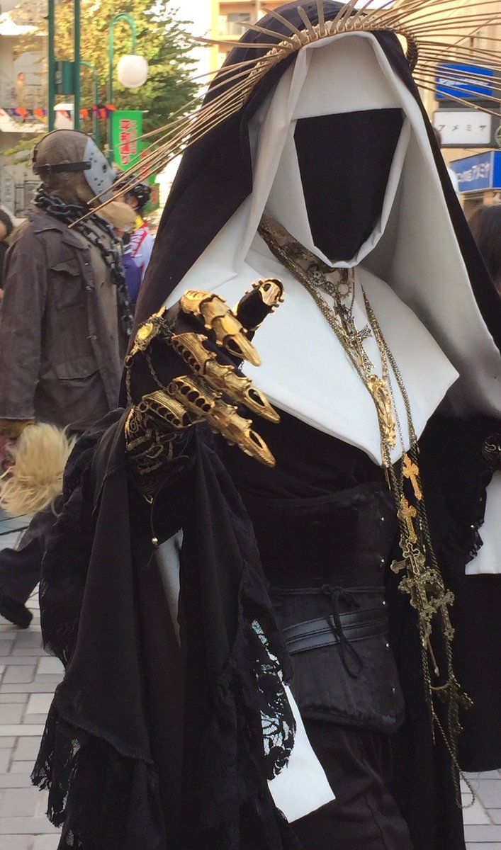 a woman dressed in a gothic nun cosplay, with a black mask over her face, and a black and white costume, adorned with gold chains and cross. the costume is very detailed and elaborate with a beautiful lace trim on the sleeves. her hand, covered in gold gloves, is raised to the viewer.