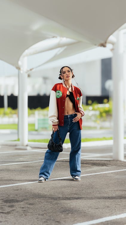 the image shows a young asian woman confidently posing in a stylish outfit in an outdoor parking lot. she's wearing a red and white varsity jacket, loosefitting blue jeans, and darkcolored sneakers. her accessories include a small black handbag.  her hair is styled up, and she carries herself with an air of confidence. the background is a modern, minimaliststyle parking structure with white canopies. the overall scene is bright, showcasing the woman's fashion and her attitude.