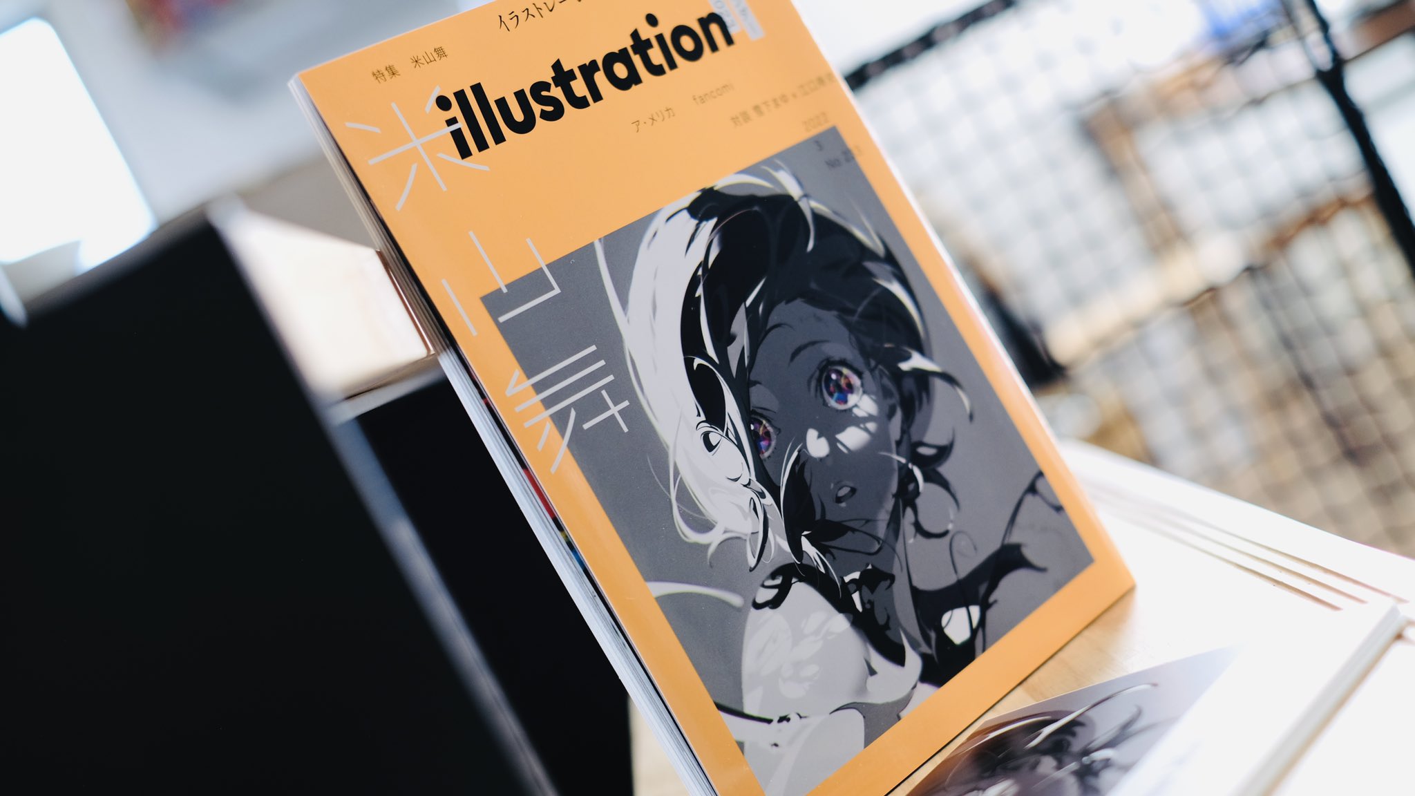 an open illustration magazine sits on a wooden surface, possibly a table or shelf. the magazine features a striking illustration of a woman with black and white hair and piercing eyes. the magazine's title, "illustration," is prominently displayed on the cover in bold, japanese characters. the background hints at a bookstore environment with shelves and other publications visible. the image is welllit, suggesting natural light streaming into the space.