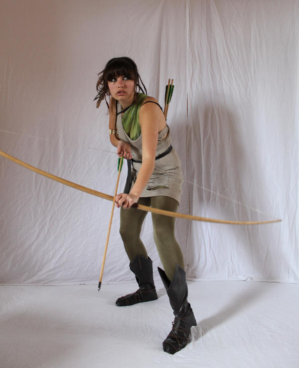 a fullbody shot of a woman dressed in a fantasy archer costume. she's holding a longbow, and her expression is one of focus and determination as she aims. the setting is a simple white backdrop, which contrasts with the details of her outfit and the wood of the bow and arrows. her stance suggests readiness, and the photograph emphasizes her skill and preparedness.