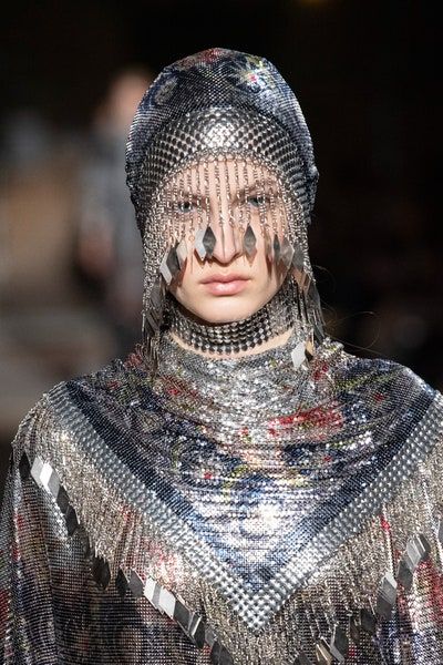 a female model wearing a silver chainmail outfit with a matching silver chainmail hat with dangling fringe that covers her face.  she also has a silver chainmail necklace.  she has a serious expression on her face and her makeup is light and natural. she is walking down a runway.