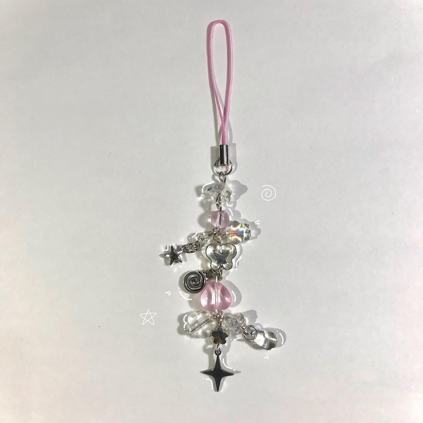 a handmade phone charm lies on a white surface. it has a pink cord attached to a silver finding, from which dangle various acrylic and silver charms, including butterflies, stars, and transparent beads.