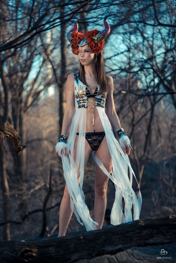 a woman in a fantasyinspired costume stands on a fallen log in a forest. she wears a horned headdress adorned with red flowers, a white and brown patterned top with a laceup front, black lace underwear, and sheer white fabric draped around her waist and legs. she has arm cuffs with fur accents and her makeup is dramatic. the setting is a natural wooded area, lit with soft, diffused light, possibly at dusk. the overall aesthetic is ethereal and mystical.