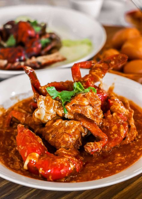 a white plate filled with chili crab. the crab is cooked in a thick red sauce and is garnished with cilantro. there is a plate of crab in the background, and a bowl with more crab meat.