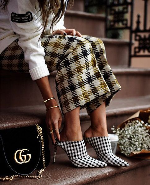 the image shows a stylish woman sitting on steps, wearing a white sweater, gingham patterned trousers, and black and white gingham heeled mules. she is also carrying a black gucci handbag with a gold chain strap.  the overall style is chic and elegant. a bouquet of baby's breath is visible next to her. the scene is outdoors, likely in a city setting.