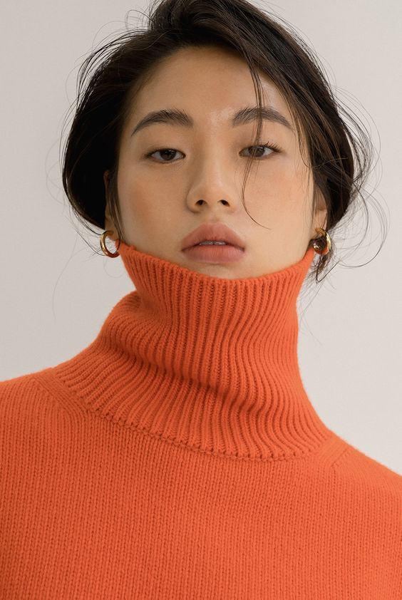 closeup portrait of a young asian woman with dark hair wearing a bright orange turtleneck sweater.  her makeup is minimal and natural, highlighting her features. she is looking directly at the camera. the overall aesthetic is minimalist and elegant.