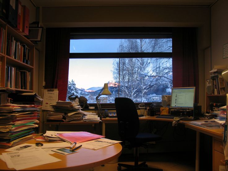 the image shows a cluttered home office with a large window overlooking a snowy winter landscape of mountains and trees.  the desk is covered in papers, files, and books, and there's a computer and chair in front of it. bookshelves are filled with books and files. a desk lamp sits on the desk, providing light. the overall feeling is one of a busy, yet cozy, workspace.