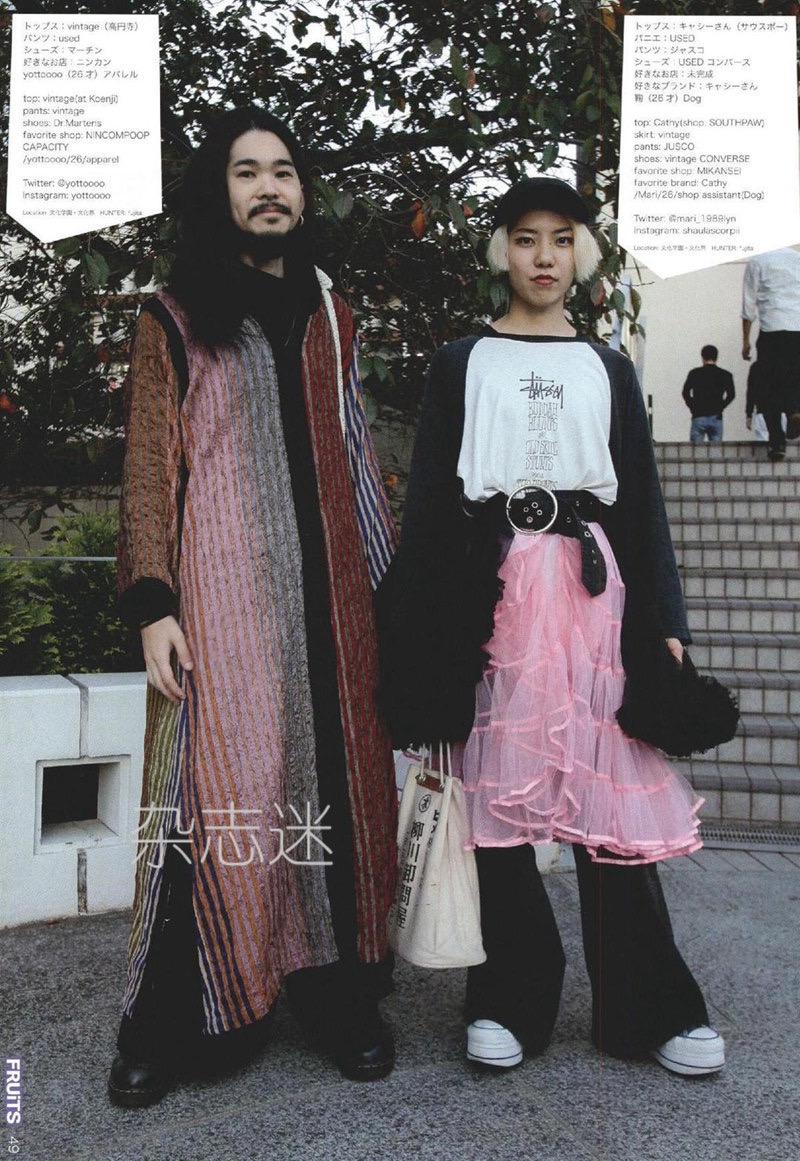 a man and a woman are posing for a fashion photo. the man is wearing a long, striped robelike garment over a black shirt. the woman is wearing a white graphic tshirt, a black cardigan, a pink tutu, and white sneakers. both are wearing backpacks. they are standing on a set of stairs in an urban environment. the photo is brightly lit with natural daylight. the overall aesthetic is eclectic and stylish, with a focus on vintage and streetwear influences. the image also contains japanese text, indicating a japanese fashion magazine or blog.