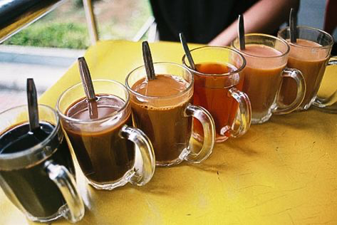 a closeup shot of six mugs of varying shades of coffee and tea sitting on a bright yellow table. the mugs are arranged in a row, with the darkest beverage on the left and the lightest on the right. each mug has a spoon resting in it and a clear glass handle. the background is blurry, suggesting an outdoor cafe or street food stall.  the drinks likely represent variations of coffee or tea based on the colors and strength. the overall mood is relaxed and casual, typical of a casual eating environment.