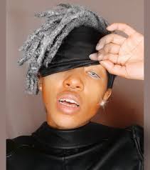 closeup portrait of a woman with grey dreadlocks wearing a black satin sleep mask over her eyes. she has on silver eyeshadow and hoop earrings, and is wearing a black turtleneck top. the lighting is soft and artificial, creating a gentle glow on her skin. the background is a neutral grey tone.