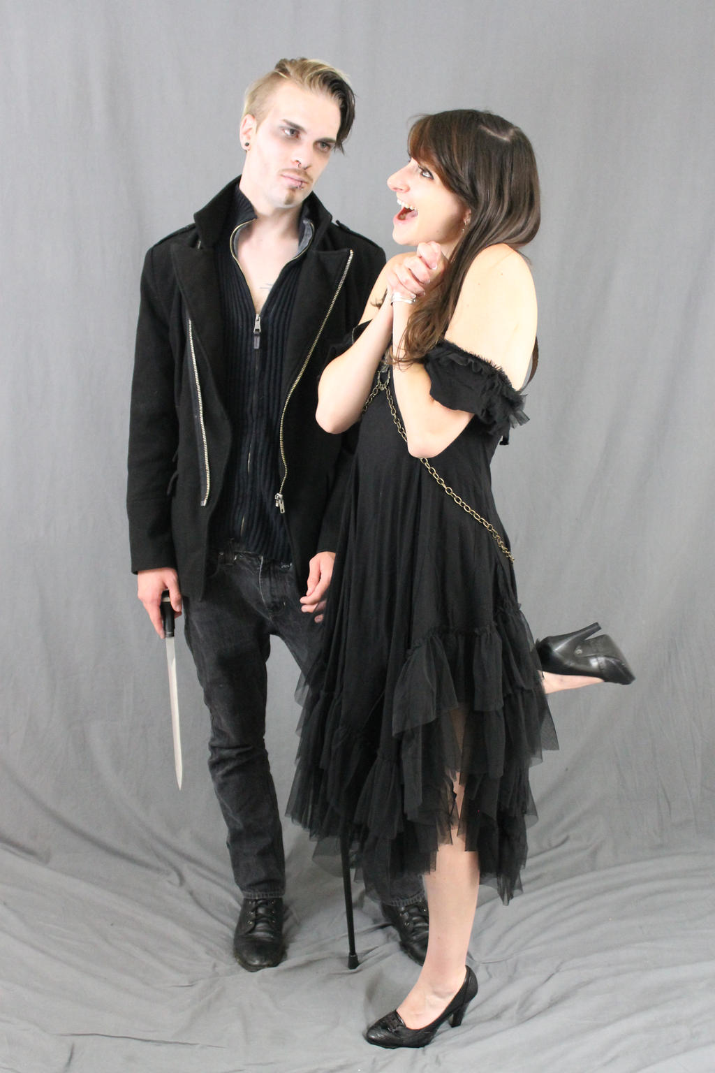 full shot of a gothic styled couple poses against a grey backdrop. the man, wearing a black jacket and dark jeans, holds a knife in his hand. the woman, in a ruffled black dress, has one arm around his neck, and is holding her other hand near her face. both are looking toward the camera, and giving a slightly distressed expression.  they wear black shoes and boots. the overall style is dark and dramatic. a cane is visible near the woman's feet.
