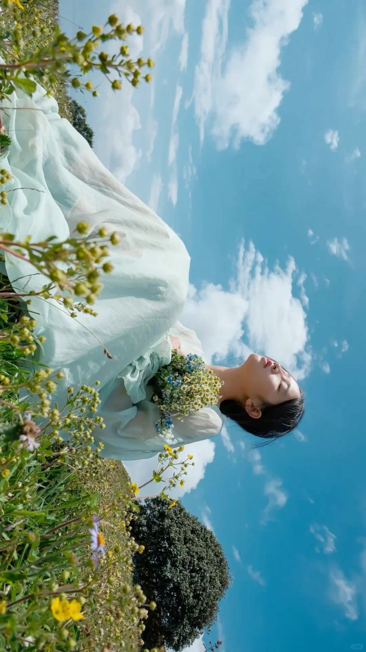 the image shows a young woman lying on her back in a field of wildflowers. she's wearing a flowing, lightgreen dress and holding a bouquet of flowers. the sky above is a bright blue with fluffy white clouds. the photograph is taken from a low angle, looking upward, creating a perspective that makes the sky seem vast and emphasizes the woman's peaceful pose. the overall mood is dreamy and serene. the flowers in the foreground and background, and the woman's dress create a harmonious color palette of greens and blues.  the slightly distorted perspective adds to the whimsical feel of the image.